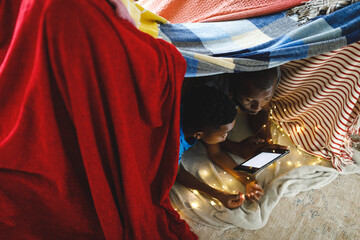 Happy african american father and son lying in blanket fort, using tablet