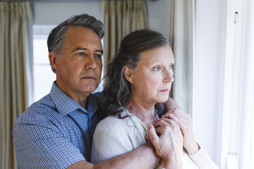 Thoughtful senior caucasian couple standing next to window, embracing