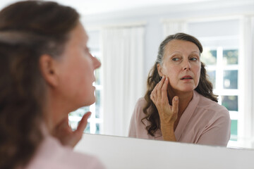 Senior caucasian woman in bathroom, looking at her face in mirror
