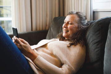 Happy senior caucasian woman in living room sitting on sofa, using smartphone