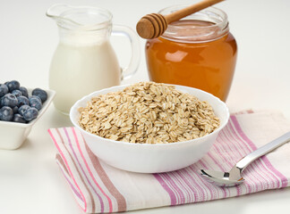 Morning breakfast, raw oatmeal flakes in a ceramic plate, milk in a decanter and honey in a jar on...