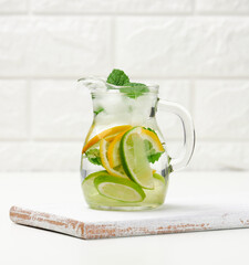 transparent glass decanter with slices of lemon, lime and mint leaves on a white table, detox