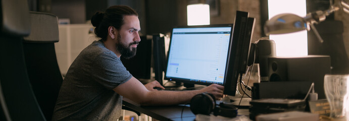 A young man plays video games in front of monitors in the evening.