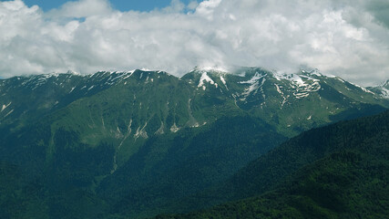View of green mountains with snowy peaks. Blue sky and clouds cover the mountain peak. Traveling and resting in the mountains in summer, hiking, climbing the mountain. Mountain landscape.