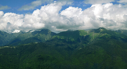 Fototapeta premium View of green mountains with snowy peaks. Blue sky and clouds cover the mountain peak. Traveling and resting in the mountains in summer, hiking, climbing the mountain. Mountain landscape.