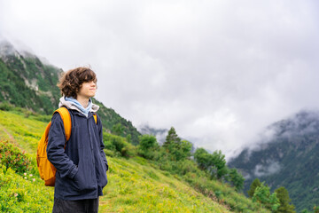 Portrait of a young teenager boy with curly hairs , with yellow backpack in a black jacket standing in the mountains in French Alps, looking away with pleasure. Hiking activities in summer.