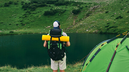 A male tourist with a backpack and a rubber mat stands near a tent by the lake. Travel, vacation, tourism.