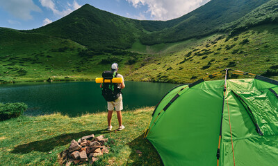 A male tourist with a backpack and a rubber mat stands near a tent by the lake. A young hiker at...