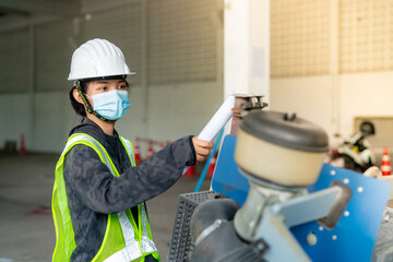 Young Asian female engineer wearing a medical mask to cover her mouth and wearing a white safety helmet Inspecting machinery in the construction zone.
