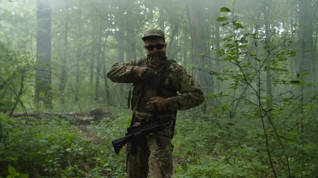 Fully equipped military man in camouflage with assault rifle walking through dense forest, observing enemy position, throwing hand grenade ahead, weapon in firing position ready to fire