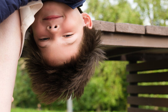 Eight-year-old boy hangs upside down from a bench at his mother's legs. Child is having fun while walking in the park. Funny picture about of children's pranks