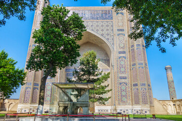 Obraz premium Facade of Bibi-Khanym mosque in Samarkand, Uzbekistan. Ahead, in shade of trees, is old stone stand for reading the Quran. Complex was built in XV century. Included in UNESCO