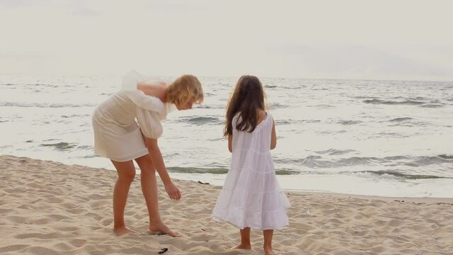 Two pretty girls in white dresses are looking for shells in the sand on the beach.