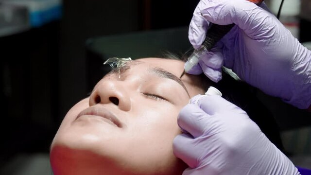 Close up microblading eyebrow tattoo. Beautician in gloves makes permanent makeup correction of the shape of the eyebrows to a young women. Eyebrow tattoo in a beauty salon.