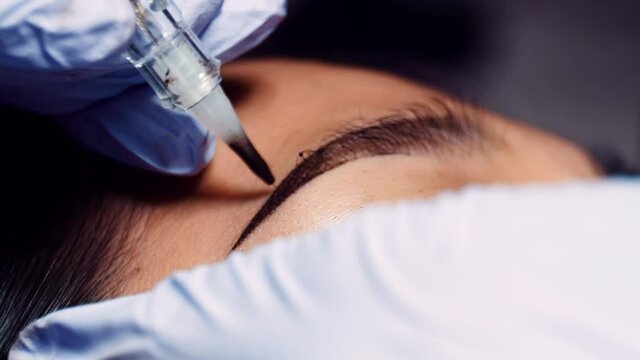 Close up microblading eyebrow tattoo. Beautician in gloves makes permanent makeup correction of the shape of the eyebrows to a young women. Eyebrow tattoo in a beauty salon.