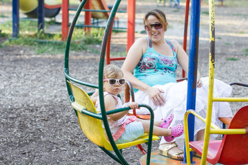 Happy mother and daughter with sunglasses fun on carousel in park on summer day. 