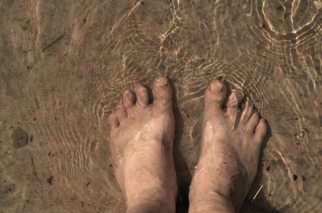 A man stands barefoot in the piercing water. A view from above of a grown man's dirty feet. Summer, daytime.