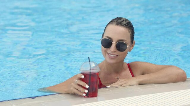 A young beautiful smiling girl is resting by the pool in a red swimsuit and sunglasses, drinking coctail. Charming millennial woman with wet blonde hair relaxing on vacation at resort. 