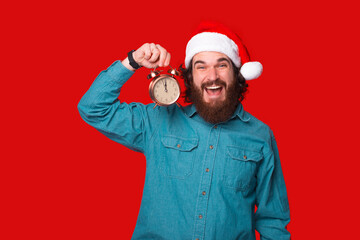 Charming young man with beard in santa claus hat holding alarm clock over red background.