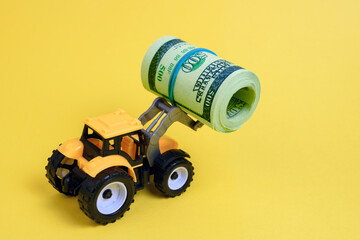 Fototapeta premium a toy bulldozer on a yellow background, holding a rolled-up bundle of dollars in a bucket , concept