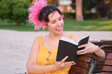Obraz premium Young caucasian woman with pink hair sitting in a bench and reading a book.