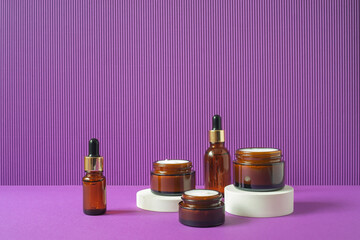 Amber glass bottles and jars with trendy geometric shapes on a violet background. White pedestals . Skincare products , natural cosmetic. Beauty concept for face and body care