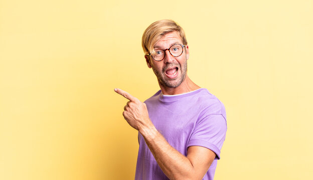 handsome blond adult man looking excited and surprised pointing to the side and upwards to copy space
