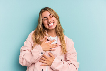 Young caucasian blonde woman isolated on blue background  laughs happily and has fun keeping hands on stomach.