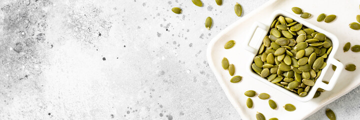 Pumpkin seeds in a white bowl on a light gray culinary background. Banner top view from the copyspace