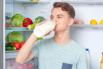 Cute young teen boy holding bottle of milk and drinks while standing near open fridge in kitchen at...