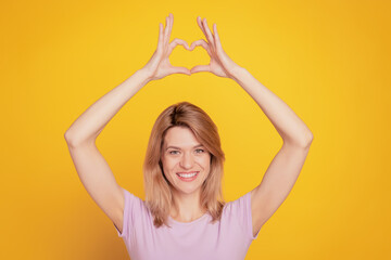 Fototapeta premium Portrait of adorable sweet lady demonstrate heart symbol raise hands on yellow background