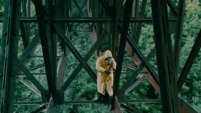 A man in a gas mask and raincoat makes a movement with his hand standing over the abyss on the floors of the iron bridge.