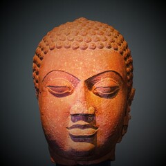 Ancient Buddha Head in state of inward-looking meditation, with half closed eyes and downward gaze, carved of red sandstone on dark gray background.