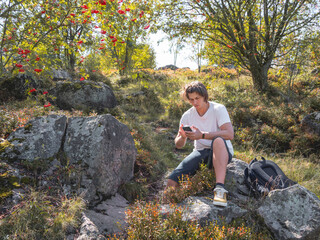 Frowning man looks at smartphone screen. Orientation on terrain using built-in navigator application. Hiking with backpack in fall season. Bright autumn leaves.