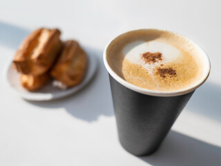 Carton cup of cappuccino coffee and plate with eclairs. Tasty dessert and hot beverage with frothy milk and cinnamon. Hard sunlight. Coffee break or breakfast.