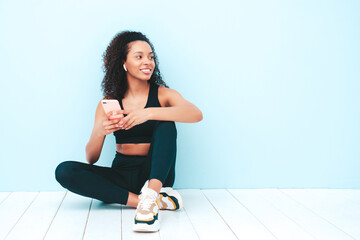 Obraz premium Fitness smiling black woman in sports clothing with afro curls hairstyle.She wears sportswear. Beautiful model with perfect tanned body.Female sitting in studio.Using apps at smartphone after training