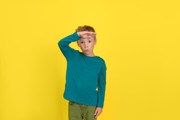 Portrait of a six year old cute male boy posing against a yellow wall in the studio, holding his palm on his forehead and looking into the distance.