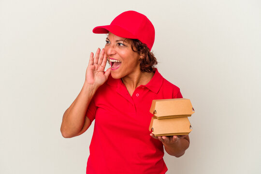 Middle age delivery woman taking burguers isolated on white background shouting and holding palm near opened mouth.