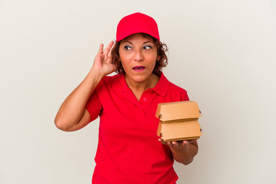 Middle age delivery woman taking burguers isolated on white background trying to listening a gossip.