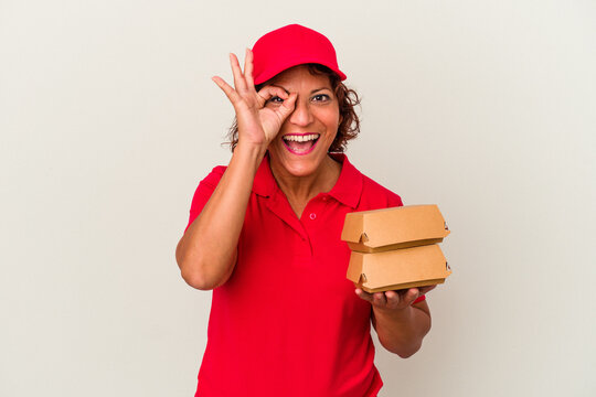 Middle age delivery woman taking burguers isolated on white background excited keeping ok gesture on eye.