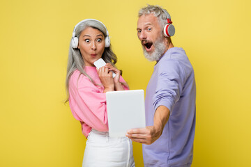 surprised middle aged and interracial couple in wireless headphones looking at digital tablet isolated on yellow