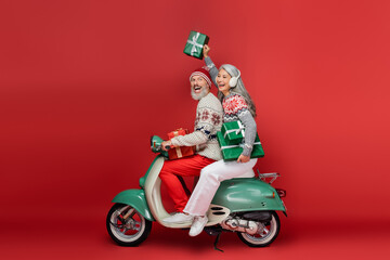 cheerful middle aged asian woman in ear muffs holding christmas presents while riding motor scooter...