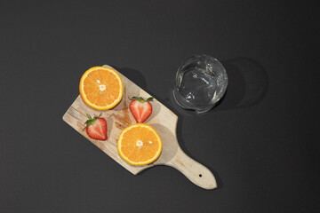 Summer scene with orange and strawberry on a wooden board with glass of water, on a black background. Minimal aesthetic.