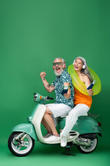 Fototapeta na wymiar multiethnic and middle aged couple in wireless headphones holding passports with air tickets and riding moped on green