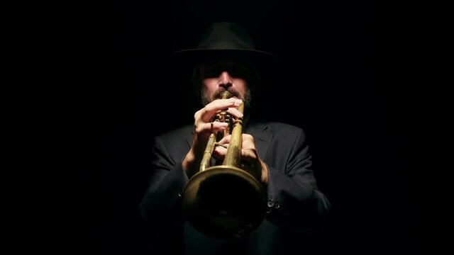 Talented male musician in hat and jacket plays musical trumpet in studio on black background, closeup, front view. Concept of instrumental music, art and jazz