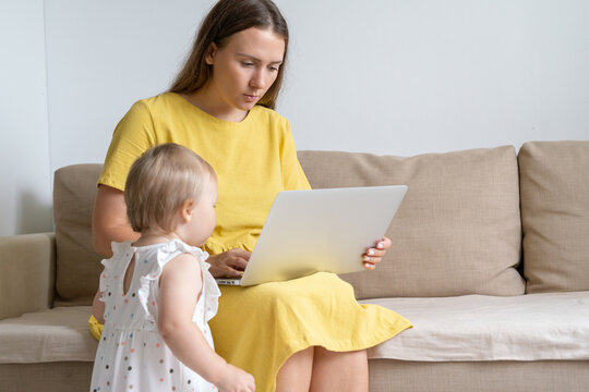 Blond Baby In Dress Standing Next To Mother Sitting On Couch And Looking At Laptop Screen. Curious Toddler Distractioning Mother From Working At Home. Concept Of Remote Work Challenges