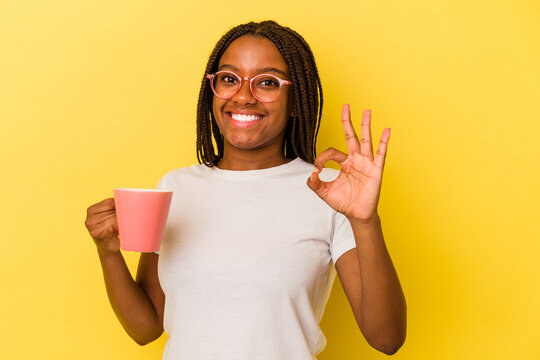 Young african american woman holding a mug isolated on yellow background  cheerful and confident showing ok gesture. - Powered by Adobe