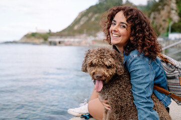 Cheerful caucasian woman hugging her water dog across the harbor walk. Horizontal view of woman traveling with animal. Animals and travel concept.