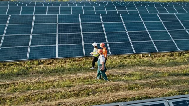 Three employees of alternative power plant walking and talking about scheme of solar panels. Technician and investor walking in Solar cell Farm through field of solar panels checking the Solar