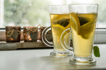 Close-up of couple of glasses on windowsill with vitamin green tea with lemon and orange. Tea bags. Candles on background.